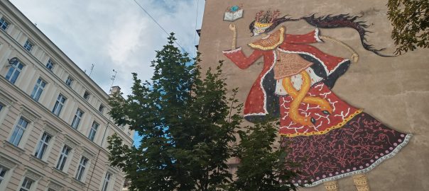 Mural przedstawiający kobietę czytającą książkę. Na pierwszym planie korona drzewa, w tle inne kamienice