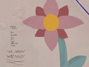 Fragment malunku na ścianie, widoczny jest kwiat. Obok niego podpisy dzieci, które stworzyły mural "nasz ogród"