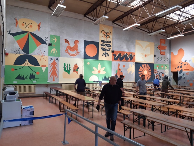Na zdjęciu ludzie rozkładający stoły w dużej sali, w tle na ścianie mural