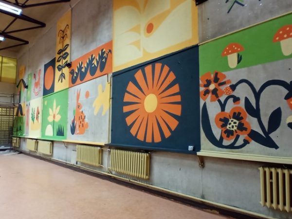 Mural na ścianie; na pierwszym planie pomarańczowe słońce, krzaki poziomek, zając; malunek w kolorach pomarańczowym, żółtym, czarnym, zielonym