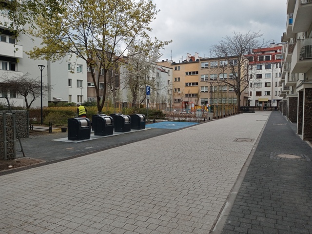 Ciąg pieszo jezdny w otoczeniu bloków z niskiej zabudowy; po lewej stronie zdjęcia kosze podziemne z widocznym wrzutami na śmieci