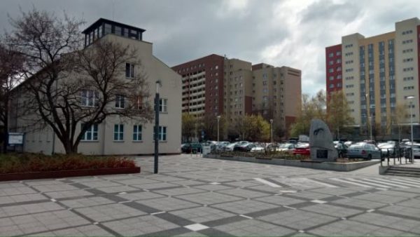 Zdjęcie przedstawiające wybrukowany plac. Istotą jest pomnik po prawej stronie w formie skalnej bryły, po lewej budynek dawnego lotniska; w tle wysokie budynki mieszkalne