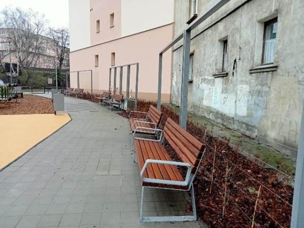 Chodnik z ławkami po prawej stronie, a za nimi metalowy trejaż ogrodowy z młodymi nasadzeniami