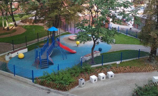Zdjęcie z góry na plac zabaw w otoczeniu zieleni. Widoczne przede wszystkim niebiesko czerwone metalowe urządzenie zabawowe z platformami i zjeżdżalniami