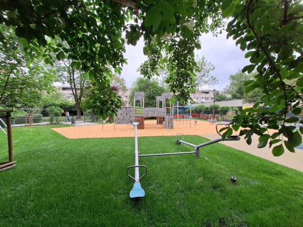 Na pierwszym planie huśtawki równoważne umiejscowione na trawie, dalej plac zabaw na drewnianych zrębkach