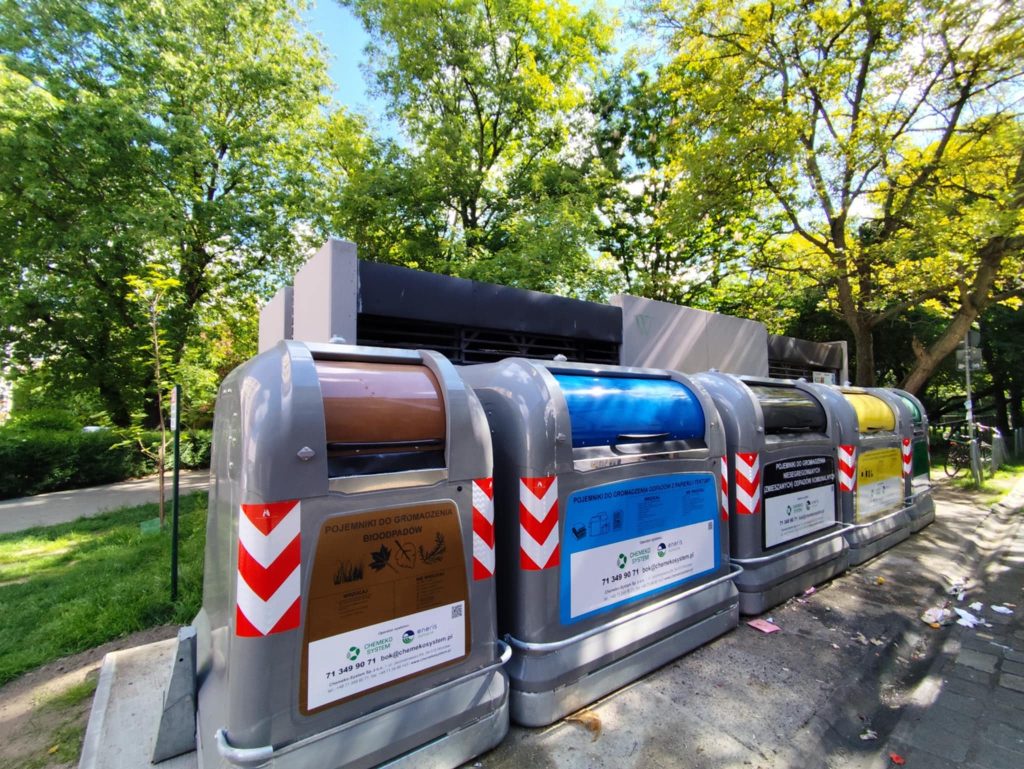 Na fotografii systemowe pojemniki na śmieci - w jednym rzędzie umieszczone są metalowy kubły na szynach z widocznym oznakowaniem frakcji