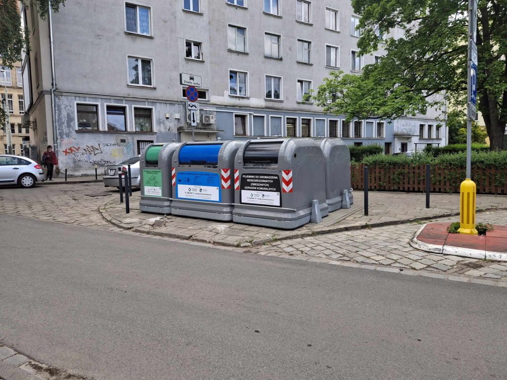 Na fotografii systemowe pojemniki na śmieci - w jednym rzędzie umieszczone są metalowy kubły na szynach z widocznym oznakowaniem frakcji
