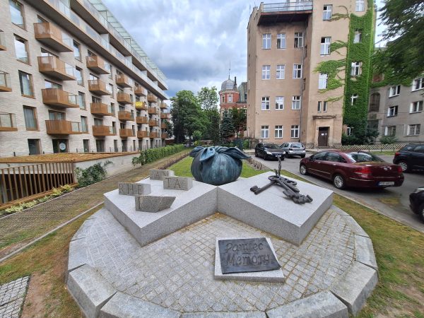 Widok na rzeźbę składającą się z granitowych bloków; na jednym kamiennie bryły, na każdej napis razem tworzący zwrot "panta rei", na drugim (środkowym) widoczny jest węzełek. A na bryle po prawej stronie umieszczone są klucze. Przed rzeźbą napis "Pamięć - memory". W tle po obu stronach fotografii budynki.