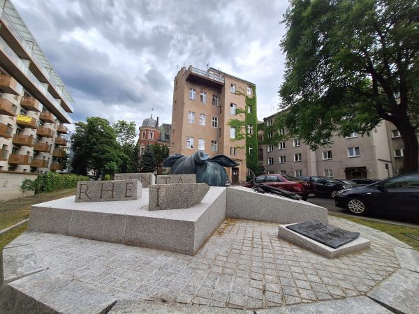 Widok na rzeźbę składającą się z granitowych bloków; na jednym kamiennie bryły, na każdej napis razem tworzący zwrot "panta rei", na drugim widoczny jest węzełek. W tle kamienica