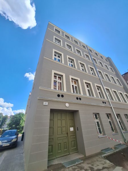 Widok od dołu na kamienicę w szarym kolorze z białym obramowaniem stolarki okiennej; na pierwszym planie zielone drzwi