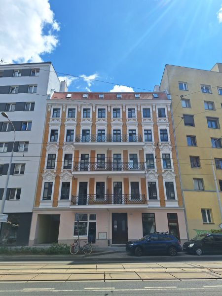 Widok na wyremontowaną fasadę kamienicy z XIX wieku; kamienica pomalowana na żółty kolor z białymi elementami; kamienica stoi w zwartej zabudowie