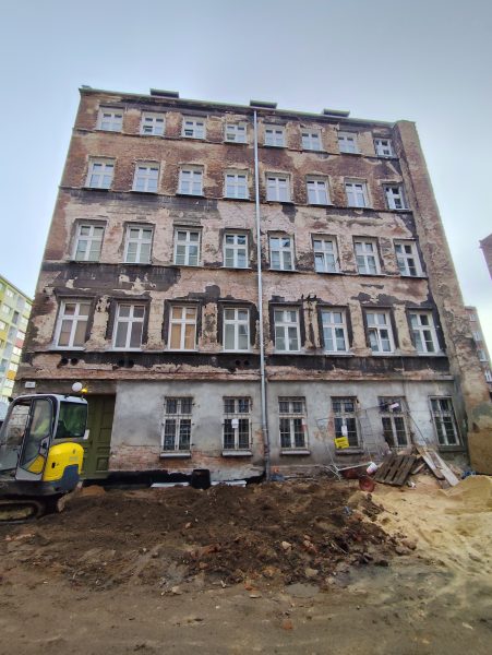 Fasada zniszczonej elewacji kamienicy z XIX wieku, po lewej stronie widoczna mała koparka