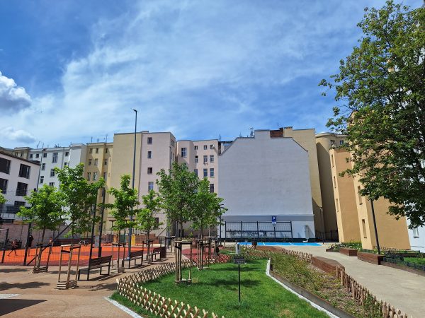 Na pierwszym planie wyremontowana część podwórka, po lewej stronie rosnące drzewa, dalej w tle kamienice