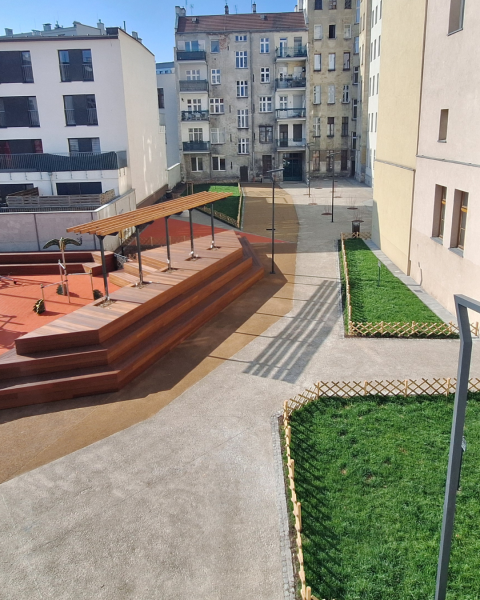 To samo miejsce, ale widok na wyremontowaną przestrzeń. Założone trawniki po prawej, sportową trybunę po lewej stronie. Wszystko w otoczeniu innych budynków.