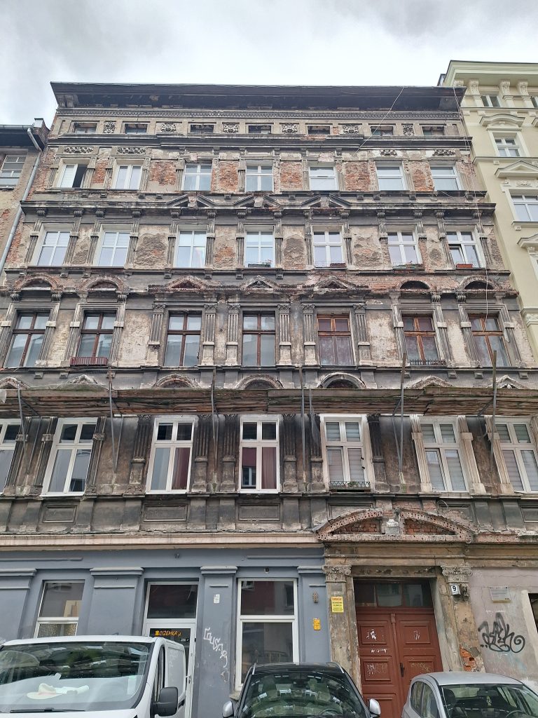 Na zdjęciu miejska kamienica bez tynki z widoczną czerwoną cegłą oraz zabepieczeniami