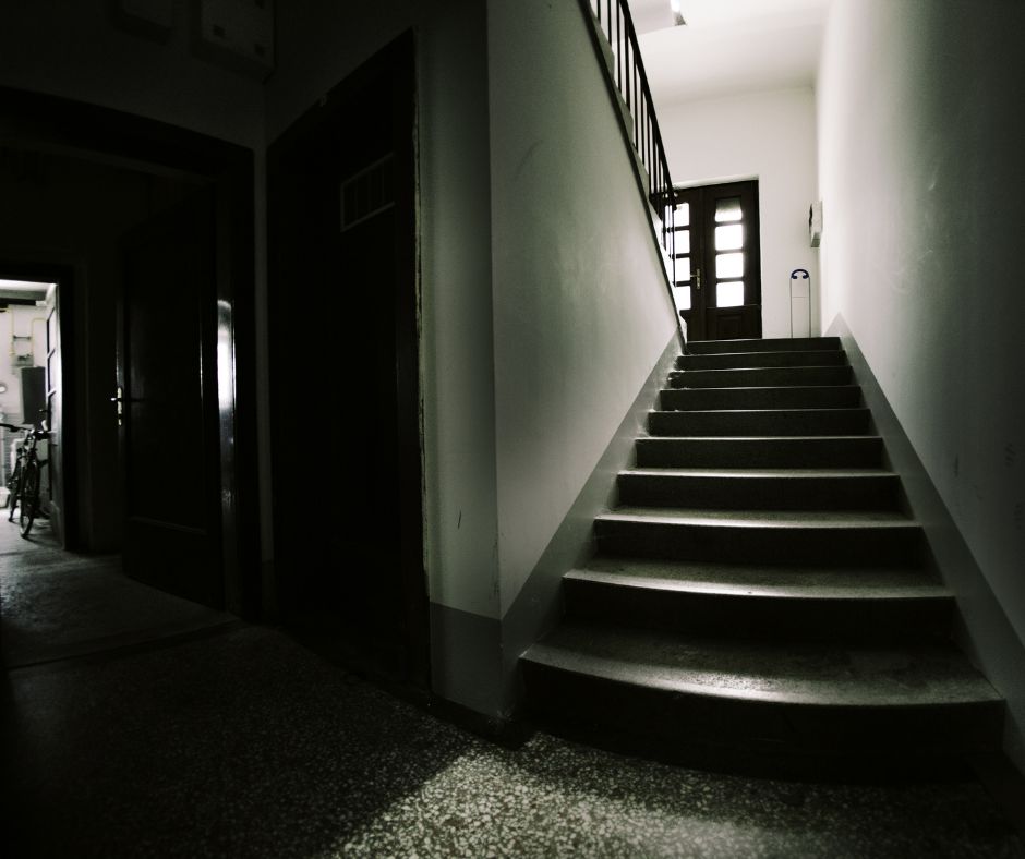 Widok na klatkę schodową. Z prawej strony od połowy zdjęcia widoczne są schody prowadzące do góry; dalej widoczne są drzwi. Lewa strona fotografii, to ciemny korytarz z lekkim światłem na końcu