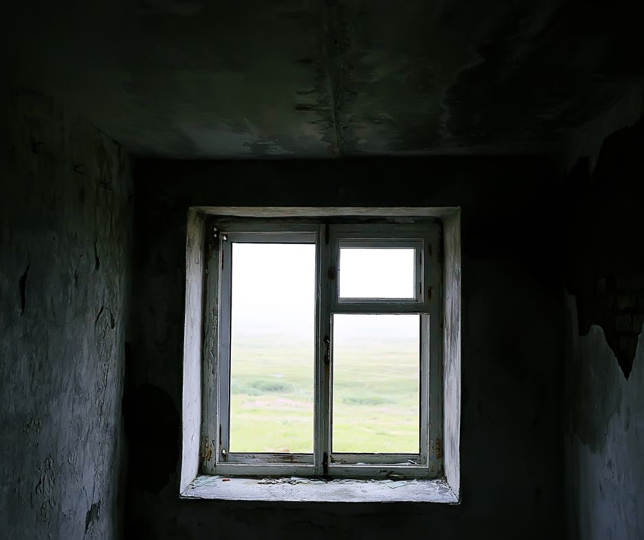 Widok na okno widziane z wnętrza lokalu. Okno bez żadnych ozdób, ciemne ściany. Można odnieść wrażenie, że lokal jest niezamieszkały