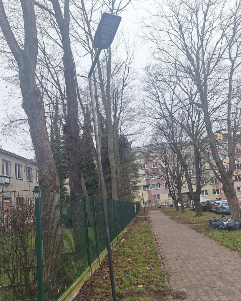 fotografia przedstawia chodnik, na którego końcu znajduje się blok mieszkalny; po lewej stronie drzewostan z widoczną lampą solarną