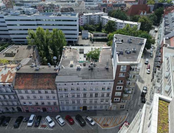 Widok z lotu ptaka na budynek. Obok stoją inne zabudowania. W dole widoczna jazdnia oraz zaparkowane auta