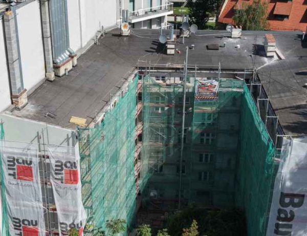 Widok z lotu ptaka na remontowany budynek. Widoczny jest płaski dach z papy. Przy elewacji stoi osiatkowane rusztowanie