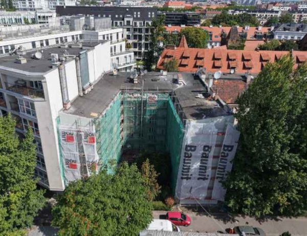 Widok z lotu ptaka na remontowany budynek. Przy elewacji stoją rusztowania, dach jest z papu, obok inne budynki. Po pokocha fotografii widoczne są zielone drzewa.