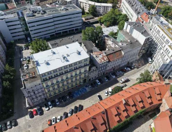 Widok z góry na ulicę wzdłuż której stoi miejska zwarta zabudowa. Po prawej stronie charakterystyczny spadzisty czerwony dach