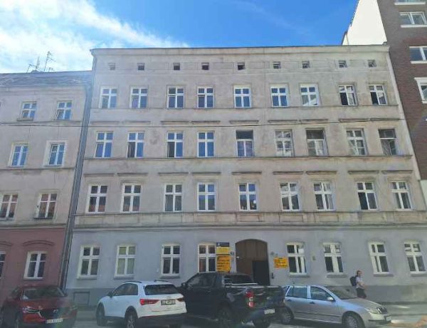 Widok na kamienice w zwartej zabudowie. Jasna elewacja, cztery kondygnacje. Przez budynkiem stoją zaparkowane auta