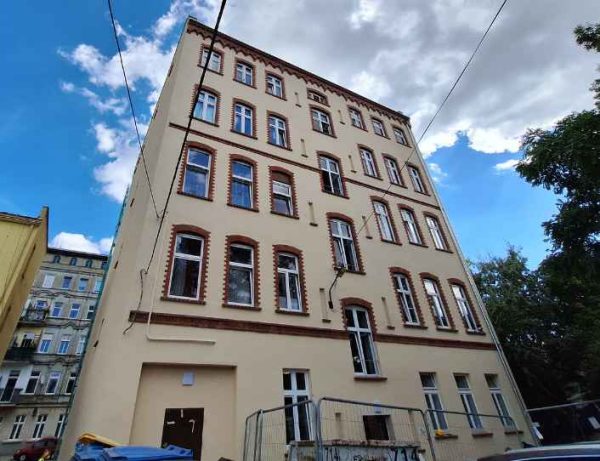 Widok z dołu na wolnostojącą kamienicę. Wyremontowany tył budynku, elewacja w jasnożółtym kolorze z mocnym akcentem wokół okien