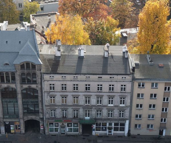 Widok z drona na pierzeję kamienic. Szczególną uwagę przyciąga środkowa, o której jest materiał. W tle drzewa jesienią.
