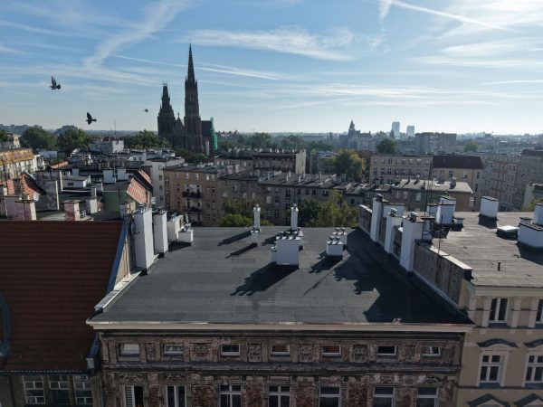 Widok z drona na płaski dach pokryty papą w zwartej pierzejowej zabudowie. W tle miejska zabudowa, widoczne wieże kościoła