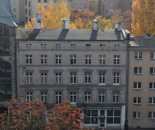 Widok z drona na pierzeję kamienic. Szczególną uwagę przyciąga środkowa, o której jest materiał. W tle drzewa jesienią.