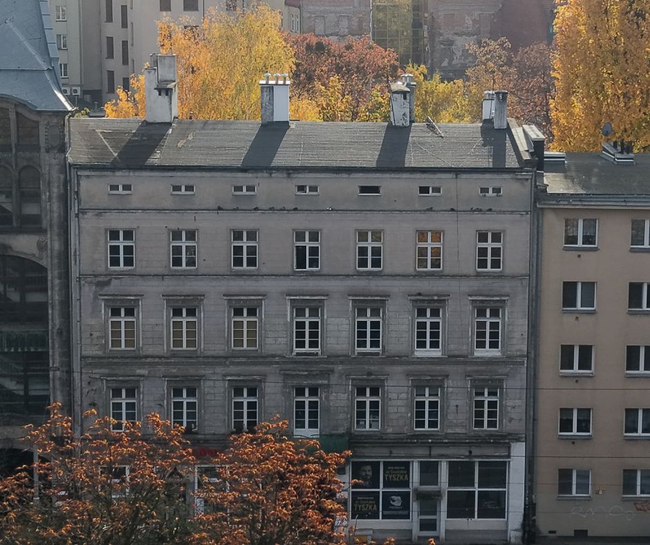 Widok z drona na pierzeję kamienic. Szczególną uwagę przyciąga środkowa, o której jest materiał. W tle drzewa jesienią.