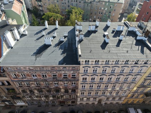 Widok z drona na płaskie dachy (dwa) pokryty papą w zwartej pierzejowej zabudowie. Widoczna jest również elewacja kamienic. Po prawej stronie, jasnożółta wyremontowana, po lewej bez zaprawy tynkarskiej.